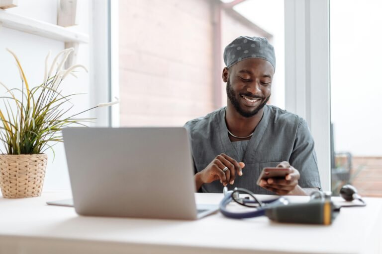 Healthcare professional smiling while using a phone beside a laptop.