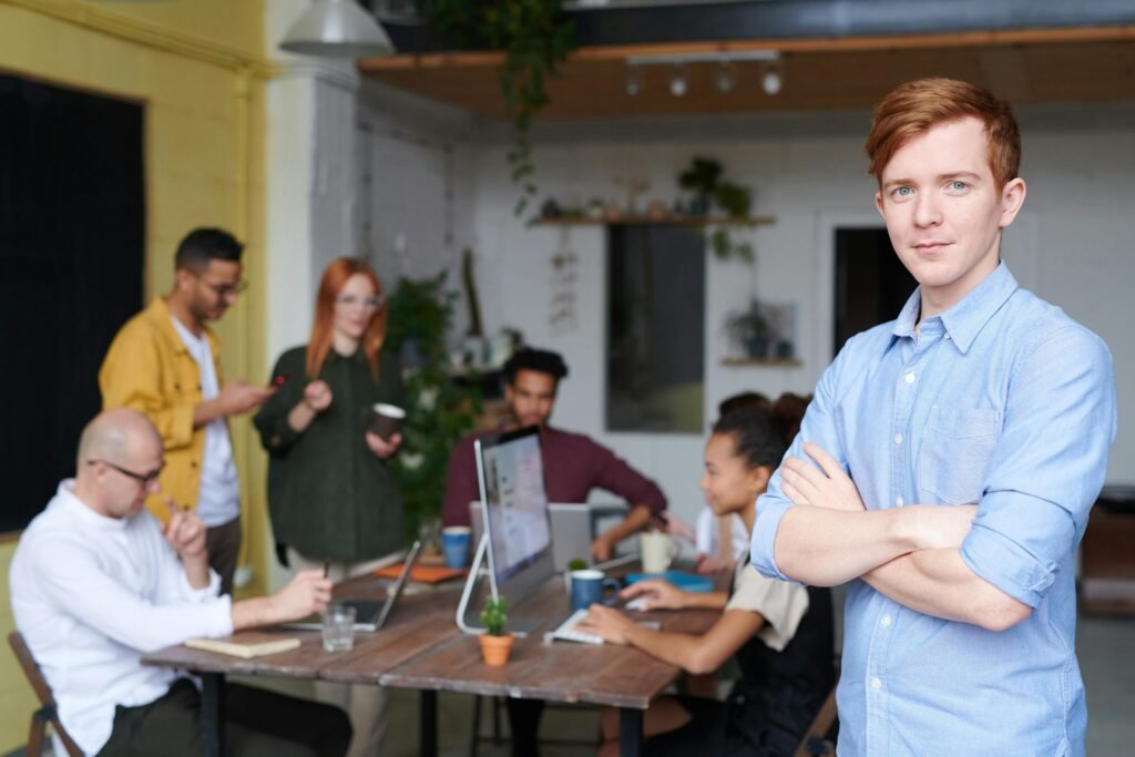 A confident young man standing with his arms crossed in the foreground, with a team collaborating in the background in a modern office.