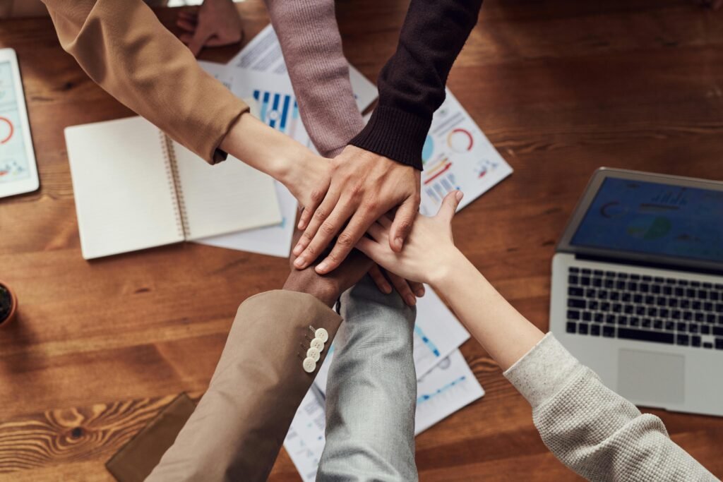 A diverse group of hands coming together in a supportive gesture over a wooden table with documents and a laptop.